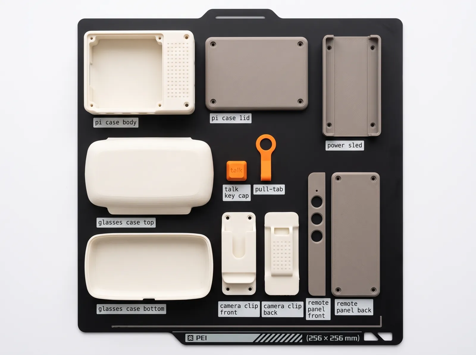 Overhead view of a black printer build plate holding every printed part laid flat, each with a small label: Pi case body and lid, power sled, glasses case top and bottom, talk key cap, pull-tab, camera clip front and back, remote panel front and back.